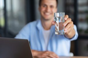 Smiling businessman or student holding glass of clean water in hand while sitting in office
